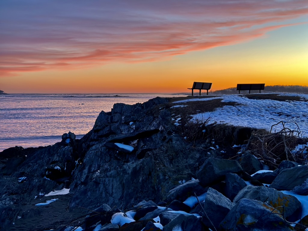 A serene coastal scene at sunset featuring two benches on a rocky outcrop overlooking the calm sea. The sky is painted with warm hues of orange and pink, reflecting on the water, with some snow visible on the ground.